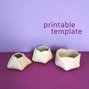 May include: Three off-white, bowl-shaped containers with folded, petal-like edges arranged in a row. The text "printable template" is displayed in purple letters above the objects. The items have a smooth, matte finish and a hollow centre.