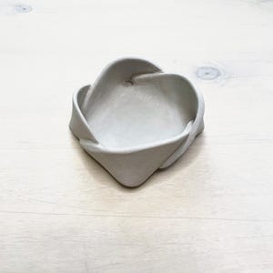 May include: A small, light gray ceramic dish with a unique, twisted design. The dish has a shallow, open center and a textured surface. The edges are folded and curved, creating an interesting shape. The dish is sitting on a light-colored wooden surface.
