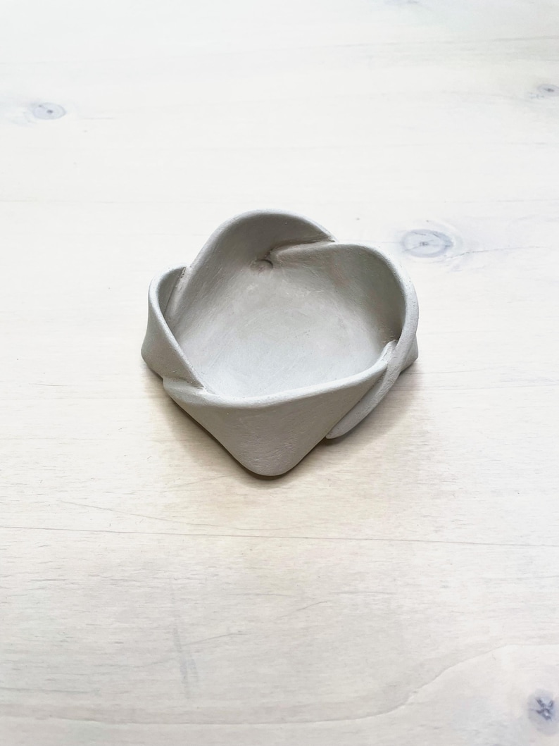 May include: A small, light gray ceramic dish with a unique, twisted design. The dish has a shallow, open center and a textured surface. The edges are folded and curved, creating an interesting shape. The dish is sitting on a light-colored wooden surface.