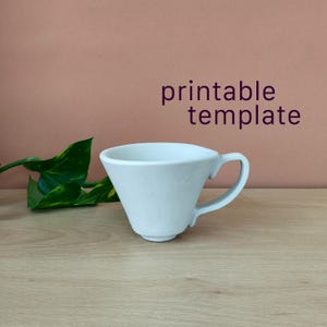 May include: A white ceramic coffee dripper with a handle, featuring a conical shape and a small base. The text 'printable template' is displayed in dark purple letters above the object, which is positioned next to a green leafy plant.