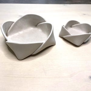 May include: Two off-white ceramic bowls with a unique folded design. The larger bowl is approximately 8 inches wide, and the smaller bowl is about 4 inches wide. The bowls have a smooth, matte finish and are suitable for decorative or functional use.