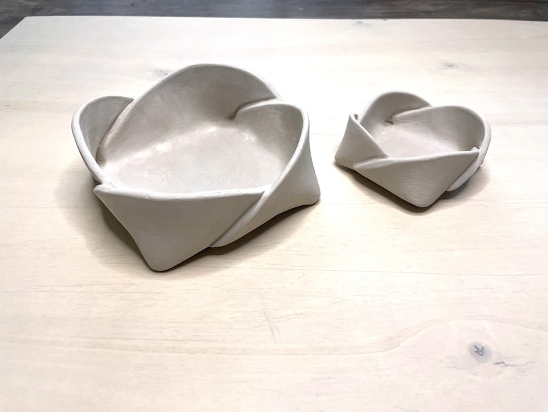 May include: Two off-white ceramic bowls with a unique folded design. The larger bowl is approximately 8 inches wide, and the smaller bowl is about 4 inches wide. The bowls have a smooth, matte finish and are suitable for decorative or functional use.
