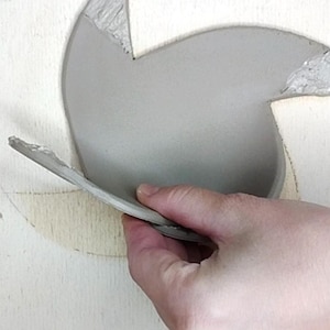 May include: A hand holds a gray, curved piece of clay, possibly part of a pottery project. The clay is smooth and light gray, with a rough edge. The background is a light-colored surface, possibly wood.