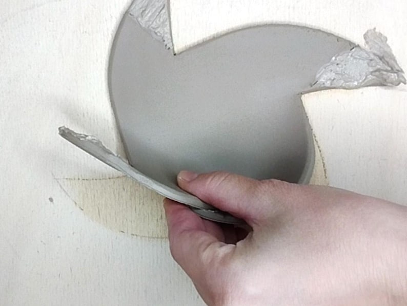 May include: A hand holds a gray, curved piece of clay, possibly part of a pottery project. The clay is smooth and light gray, with a rough edge. The background is a light-colored surface, possibly wood.