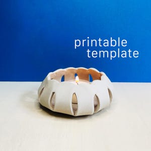 May include: A light-colored, circular candle holder with a scalloped, cut-out design holding a lit tea light candle. The text "printable template" is displayed in white letters above the object against a solid blue background.