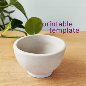 May include: A small, matte white ceramic bowl with a rounded shape and a raised base. The text "printable template" is displayed in purple letters above the bowl. The bowl has a smooth, uniform finish and a wide, open top opening.