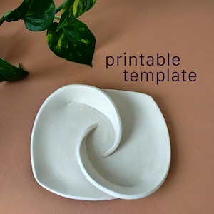 May include: A white ceramic dish with a curved, spiral-shaped divider creating two distinct sections. The text "printable template" is printed in dark purple letters above the dish. A green and yellow leafed plant is visible in the corner.