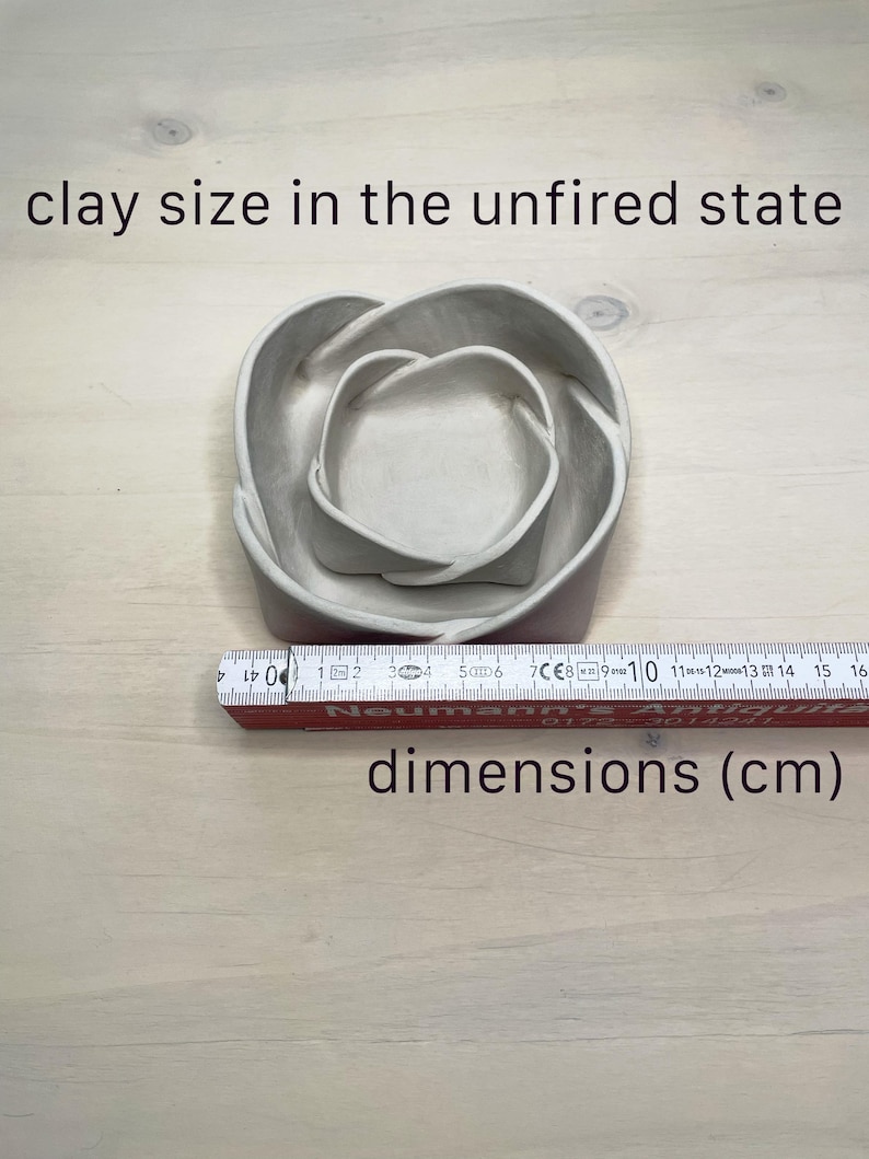 May include: A nested, unfired clay bowl set. The set includes two bowls, one inside the other, with a heart-shaped design. The text "clay size in the unfired state" and "dimensions (cm)" are visible. A ruler is placed next to the bowls.