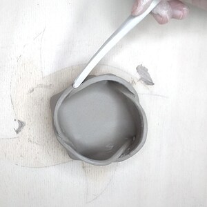 May include: A gray, handmade clay bowl with a unique, organic shape. A white tool is used to refine the rim of the bowl. The bowl sits on a light-colored surface, suggesting a pottery studio setting.