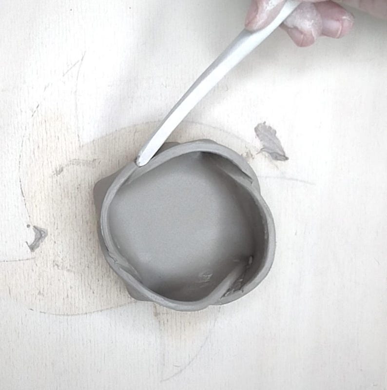 May include: A gray, handmade clay bowl with a unique, organic shape. A white tool is used to refine the rim of the bowl. The bowl sits on a light-colored surface, suggesting a pottery studio setting.