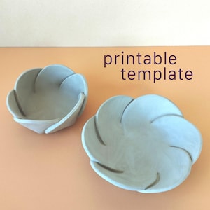 May include: Two light blue ceramic-style bowls with a layered petal design. The text "printable template" is displayed in dark purple letters above the bowls. The objects have a smooth, matte finish and a rounded, flower-like shape.