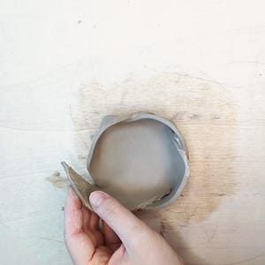 May include: A gray ceramic bowl with a partially detached rim. The bowl is held by a hand, revealing the interior. The background is a light-colored wooden surface.