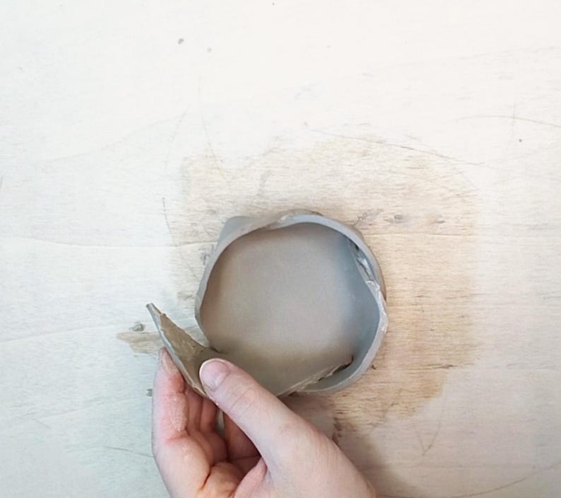 May include: A gray ceramic bowl with a partially detached rim. The bowl is held by a hand, revealing the interior. The background is a light-colored wooden surface.