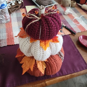 May include: A stack of three crocheted pumpkins decorated with fall leaves and orange beads. The pumpkins are burgundy, cream, and orange.