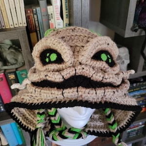 May include: A crocheted hat in tan, black, and green, resembling a character from a movie. The hat features large, green-eyed details, a black mouth, and braided accents with green and black strands. The hat is displayed on a white mannequin head.