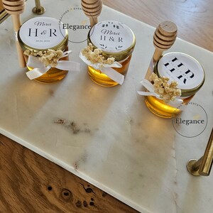 Può includere: Tre piccoli vasetti di vetro pieni di miele, ognuno con un mestolo di miele in legno e un'etichetta bianca con il testo "Merci H & R" e un motivo floreale. I vasetti sono su una superficie di marmo bianco.