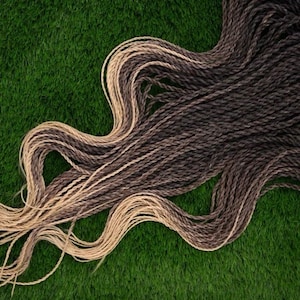 Könnte beinhalten: Geflochtene Haarverlängerungen in Braun- und Blondtönen, angeordnet in einem wellenförmigen Muster vor einem grünen Hintergrund. Das Haar ist mit einem braunen Band zusammengebunden. Die Extensions haben ein natürliches, strukturiertes Aussehen.