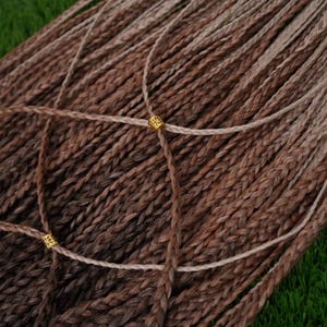 Könnte beinhalten: Nahaufnahme von braunen und hellbraunen geflochtenen Haarverlängerungen. Die Extensions sind in einem Bündel angeordnet, wobei einige Strähnen ein dekoratives Muster bilden. Kleine goldene Perlen sind an den Kreuzungen der Strähnen platziert. Der Hintergrund ist grün.