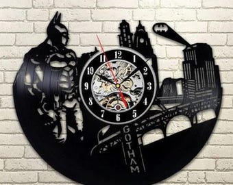 Könnte beinhalten: Schwarze Batman-Wanduhr. Die Uhr zeigt eine Silhouette von Batman, die Skyline von Gotham City und ein Fledermaussignal. Die Zeiger sind rot. Das Wort "Gotham" ist auf der Uhr zu sehen.