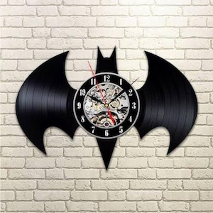 Puede incluir: Reloj de pared negro con forma de Batman. El reloj está hecho de un disco de vinilo reutilizado. La esfera del reloj es negra con números blancos y engranajes visibles. El segundero es rojo. El reloj está montado en una pared de ladrillo blanco.