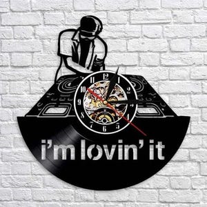 May include: Black vinyl record clock featuring a DJ silhouette and the phrase "i'm lovin' it." The clock face has a white border with black numbers and a red second hand. The clock is mounted on a white brick wall.
