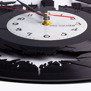 K&ouml;nnte beinhalten: Schwarze Vinyl-Schallplattenuhr mit einer Skyline-Silhouette am &auml;u&szlig;eren Rand. Das Zifferblatt ist wei&szlig; mit schwarzen Ziffern und Zeigern sowie einem roten Sekundenzeiger. Der Text "VINYL CLOCK ART" ist auf dem Zifferblatt aufgedruckt.