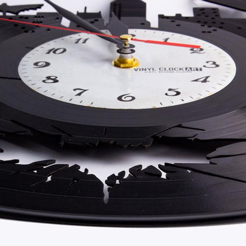 K&ouml;nnte beinhalten: Schwarze Vinyl-Schallplattenuhr mit einer Skyline-Silhouette am &auml;u&szlig;eren Rand. Das Zifferblatt ist wei&szlig; mit schwarzen Ziffern und Zeigern sowie einem roten Sekundenzeiger. Der Text "VINYL CLOCK ART" ist auf dem Zifferblatt aufgedruckt.