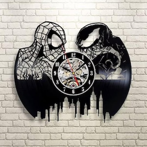 Pode incluir: Relógio de disco de vinil preto com silhuetas de Homem-Aranha e Venom em ambos os lados de um mostrador. O mostrador tem um design a preto e branco com engrenagens visíveis e ponteiros vermelhos. O relógio está montado numa parede de tijolo.