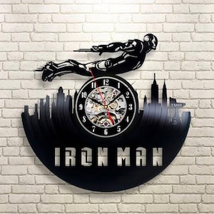 Può includere: Orologio da parete nero di Iron Man realizzato con un disco in vinile. L'orologio presenta una silhouette di Iron Man che vola sopra un paesaggio urbano e le parole "IRON MAN". Le lancette dell'orologio sono rosse e nere.