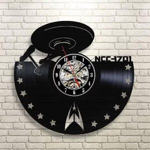 May include: Black wall clock with a Star Trek theme. The clock is made from a vinyl record and features a ship design, the text "NCC-1701", and a clock face with visible gears. The clock has a black background with white accents and star cutouts.