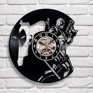 Op de afbeelding: Een zwarte vinylplaatklok met een Deadpool-ontwerp. De klok toont Deadpool in een dynamische pose, met een pistool. De wijzerplaat is zichtbaar, met een skeletontwerp in het midden. De achtergrond is een witte bakstenen muur.