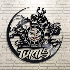 May include: Black wall clock featuring the Teenage Mutant Ninja Turtles. The clock has a black silhouette design of the turtles and the words "TEENAGE MUTANT NINJA TURTLES". The clock face is visible with black numbers and red hands.