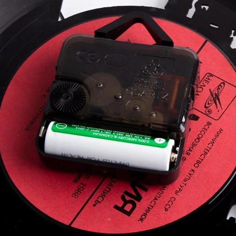 K&ouml;nnte beinhalten: Ein schwarzes Quarz-Uhrwerk mit Batteriefach, montiert auf einer roten Schallplatte. Die Uhr hat ein schwarzes Kunststoffgeh&auml;use und ein kleines Zahnrad. Die Schallplatte hat kyrillischen Text und ein Logo.