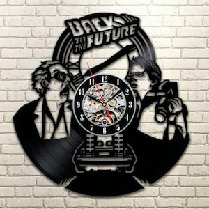 Può includere: Orologio da parete nero con un design del film "Ritorno al futuro". Il quadrante dell'orologio è in stile analogico classico con ingranaggi visibili. L'orologio è montato su un muro di mattoni.