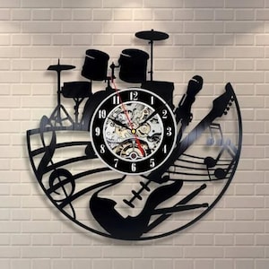 Op de afbeelding: Zwarte wandklok met een muzikaal thema. De klok heeft een drumstel, gitaar, microfoon en muzieknoten. De wijzerplaat is zwart met witte cijfers en een zichtbaar uurwerk.
