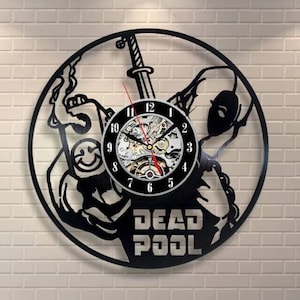 Może przedstawiać: Czarny zegar ścienny z motywem Deadpool. Zegar ma okrągłą tarczę z widocznymi kołami zębatymi i czarną ramą. Słowa "DEAD POOL" są wycięte na dole. Wskazówki zegara są czerwone i czarne.