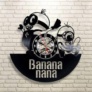 Pode incluir: Relógio de disco de vinil preto com um design de desenho animado de dois personagens com olhos grandes. O mostrador do relógio é visível, com uma moldura preta e números brancos. As palavras "Banana Nana" são exibidas abaixo.