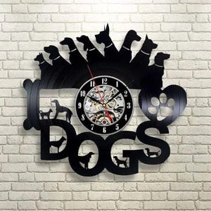 Könnte beinhalten: Schwarze Wanduhr mit Hundemotiv. Die Uhr hat ein kreisförmiges Zifferblatt mit sichtbaren Zahnrädern, umgeben vom Wort "DOGS" und Hundesilhouetten. Eine Pfotenabdruckform ist ebenfalls enthalten.