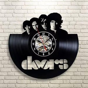 May include: Black vinyl record clock featuring a silhouette of The Doors band members. The clock face has a visible mechanical movement and white numerals. The band's name, "doors", is cut out at the bottom of the record.