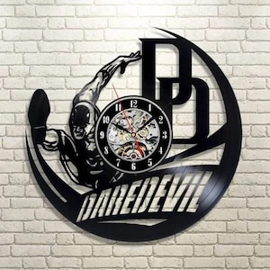 Può includere: Orologio da parete nero con la silhouette di un supereroe, la scritta "DAREDEVIL" e le iniziali "DD". Il quadrante dell'orologio ha un movimento meccanico visibile con lancette nere e una lancetta dei secondi rossa. L'orologio è montato su un muro di mattoni.