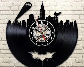 Könnte beinhalten: Schwarze Vinyl-Schallplattenuhr mit Batman-Motiv, die eine Stadtsilhouette zeigt. Das Zifferblatt hat einen weißen Rand mit schwarzen Ziffern und sichtbaren Zahnrädern. Ein Fledermaussignal und eine Batman-Figur sind ebenfalls enthalten.