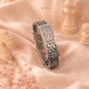 May include: A silver-toned metal bracelet featuring a central rectangular panel with a woven grid pattern, flanked by sections of intricate braided chain links. The piece has a polished finish with darkened recessed areas to highlight the texture.