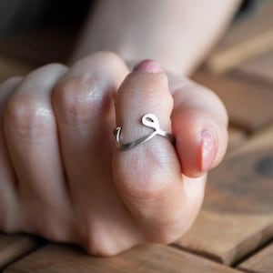 May include: A silver ring with a cursive letter 'L' design. The ring is worn on a person's finger.