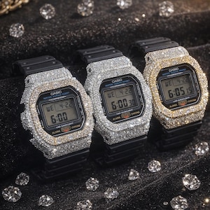 Könnte beinhalten: Drei Casio Digitaluhren, besetzt mit glitzernden Edelsteinen. Die Uhren haben schwarze Armbänder und zeigen Uhrzeit und Datum an. Eine Uhr hat eine goldfarbene Lünette, die anderen silberfarbene. Sie liegen auf dunklem Hintergrund.