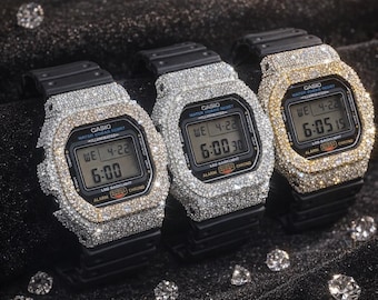 May include: Three Casio digital watches encrusted with sparkling gemstones. The watches have black bands and display the time and date. One watch has a gold-colored bezel, while the other two have silver-colored bezels. The watches are set against a dark background.