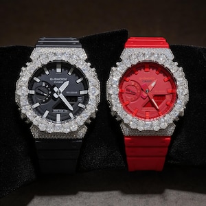 May include: Two G-Shock Casio watches with diamond-encrusted bezels. One watch has a black face and band, the other has a red face and band. Both watches have a digital display and analog hands. The watches are displayed on a black surface.