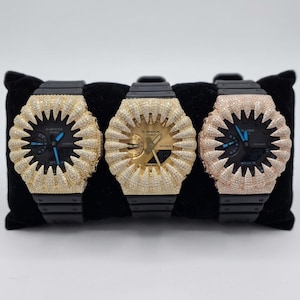 May include: Three G-Shock watches with black bands and faces. The watch bezels are encrusted with sparkling stones in gold, gold, and rose gold colors. The watches are displayed on a black velvet surface.