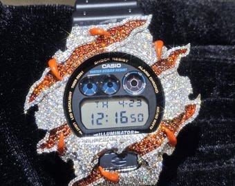 May include: Black Casio digital watch with a custom bezel encrusted with sparkling crystals and orange accents. The watch face displays the time, date, and day. The bezel design extends beyond the watch case.