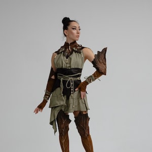 Puede incluir: Un disfraz de fantasía con un vestido verde con detalles de hojas, cubiertas de cuero marrón para brazos y piernas y un cinturón negro. El atuendo incluye un cuello alto y cuentas decorativas, creando una estética de bosque o elfo.