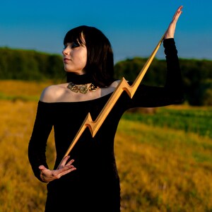 May include: A wooden lightning bolt prop with a gold finish. The prop is held up against a backdrop of a field and blue sky. The lightning bolt is approximately 30 inches long.
